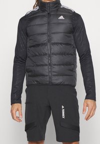 Svart pufferväst med hög krage, dragkedjestängning och horisontellt quiltat mönster. Har vitt Adidas-logotyp och randiga detaljer på axlarna.