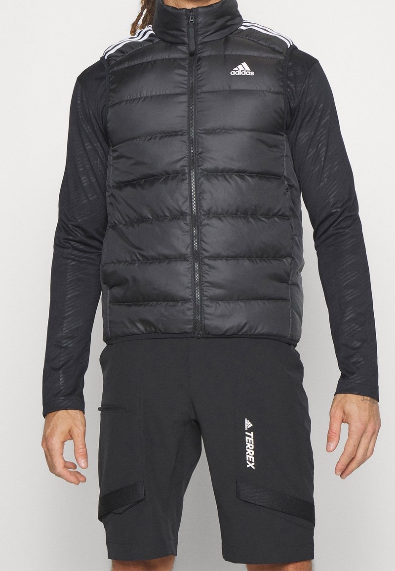 Svart pufferväst med hög krage, dragkedjestängning och horisontellt quiltat mönster. Har vitt Adidas-logotyp och randiga detaljer på axlarna.