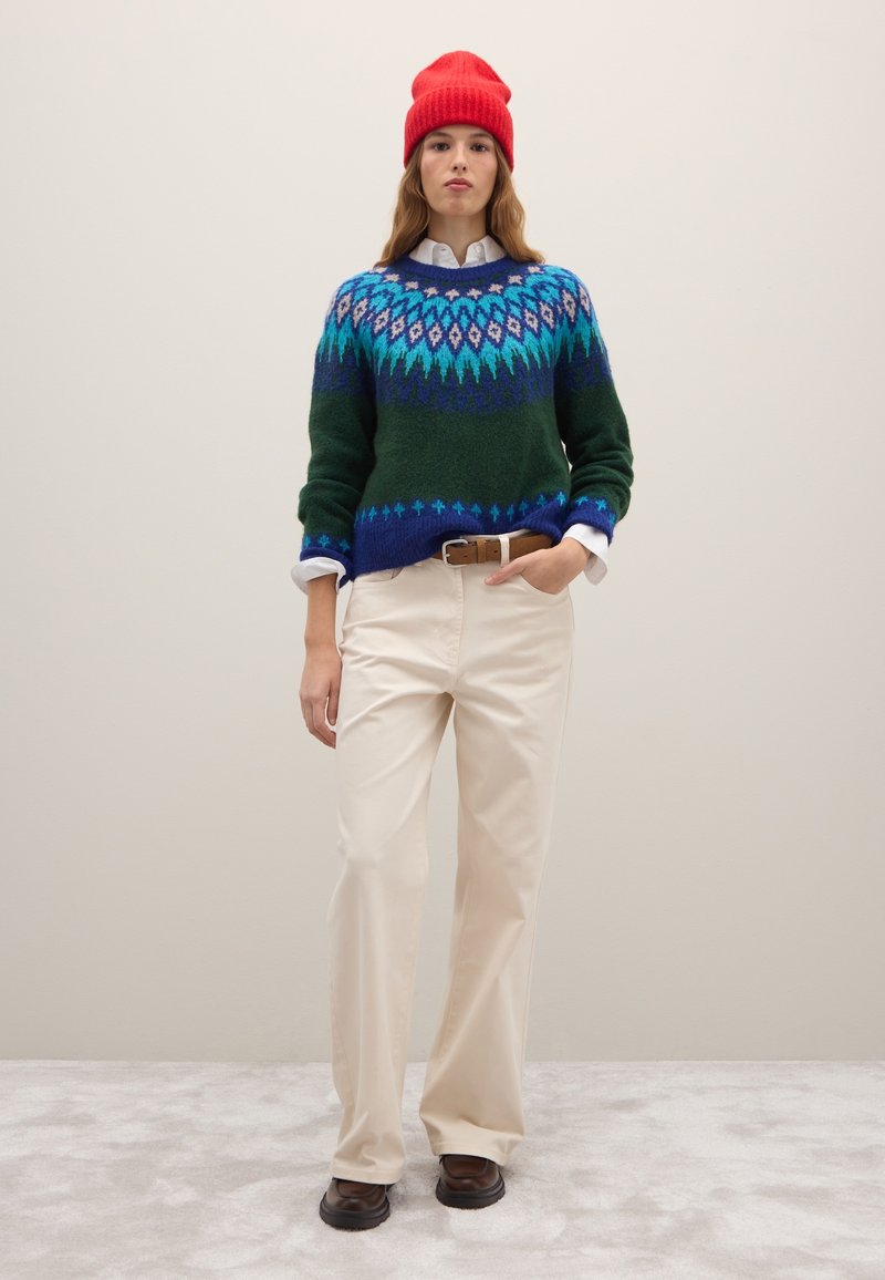 Maglione lavorato a maglia verde con motivo geometrico blu e turchese, abbinato a pantaloni crema e scarpe marroni. Berretto rosso, camicia bianca sotto.
