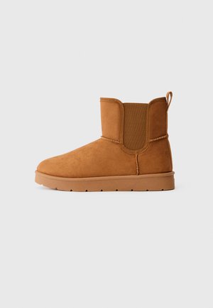 Brune ankelstøvletter i semsket skinn med elastisk sidepanel og trekkrem, flate såler og minimalistiske sømdetaljer på en lys, ensfarget bakgrunn.