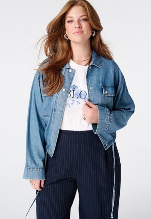 Vrouw met lang rood haar, zilveren oorringen, een spijkerjack over een wit shirt met bloemenprint en marineblauwe broek met krijtstrepen.