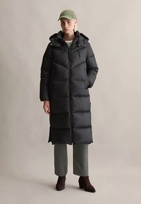 Vrouw in een lange zwarte pufferjas met capuchon, een grijs geblokte cropped broek, donkere enkellaarsjes en een lichtgroene baseballpet.