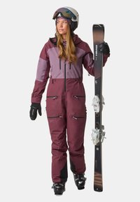 Skikleding met een bordeauxrode en paarse tweekleurige ontwerp, met ritszakken, handschoenen en een witte helm, terwijl hij een zwarte ski vasthoudt.