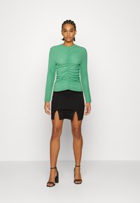 Camisola verde de mangas compridas com textura franzida na parte da frente, combinada com uma saia mini preta com fendas laterais. Modelo usa sapatos de salto pretos.