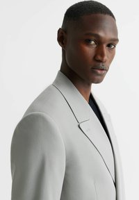 Jasnoszary blazer o strukturalnym designie, z wyciętymi klapami i gładką fakturą. Model ubrany w czarną koszulę pod spodem, zwrócony nieco w lewo.