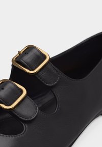 Μαύρο δερμάτινο παπούτσι με δύο χρυσές τετράγωνες αγκράφες και κοψίματα σε λευκό φόντο.