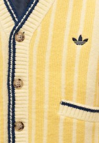 Κίτρινη και κρεμ ριγέ ζακέτα κατασκευασμένη από υφή υφάσματος. Διαθέτει ριμπ τελειώματα, τέσσερα κουμπιά, ένα ναυτικό μπλε στοιχείο και ένα κεντημένο λογότυπο.