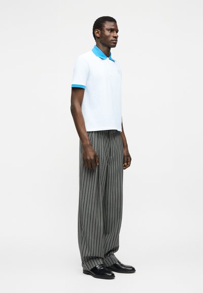 Ανδρας στέκεται φορώντας λευκό πόλο με μπλε γιακά και μανσέτες, χαλαρά μαύρα και γκρι παντελόνια με κάθετες ρίγες και μαύρα παπούτσια, με λευκό φόντο.