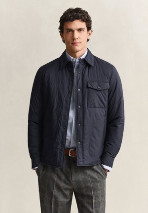 Uomo che indossa una giacca blu navy sopra una camicia azzurro chiaro con bottoni, pantaloni grigi a quadri e cintura marrone, in piedi davanti a uno sfondo neutro.