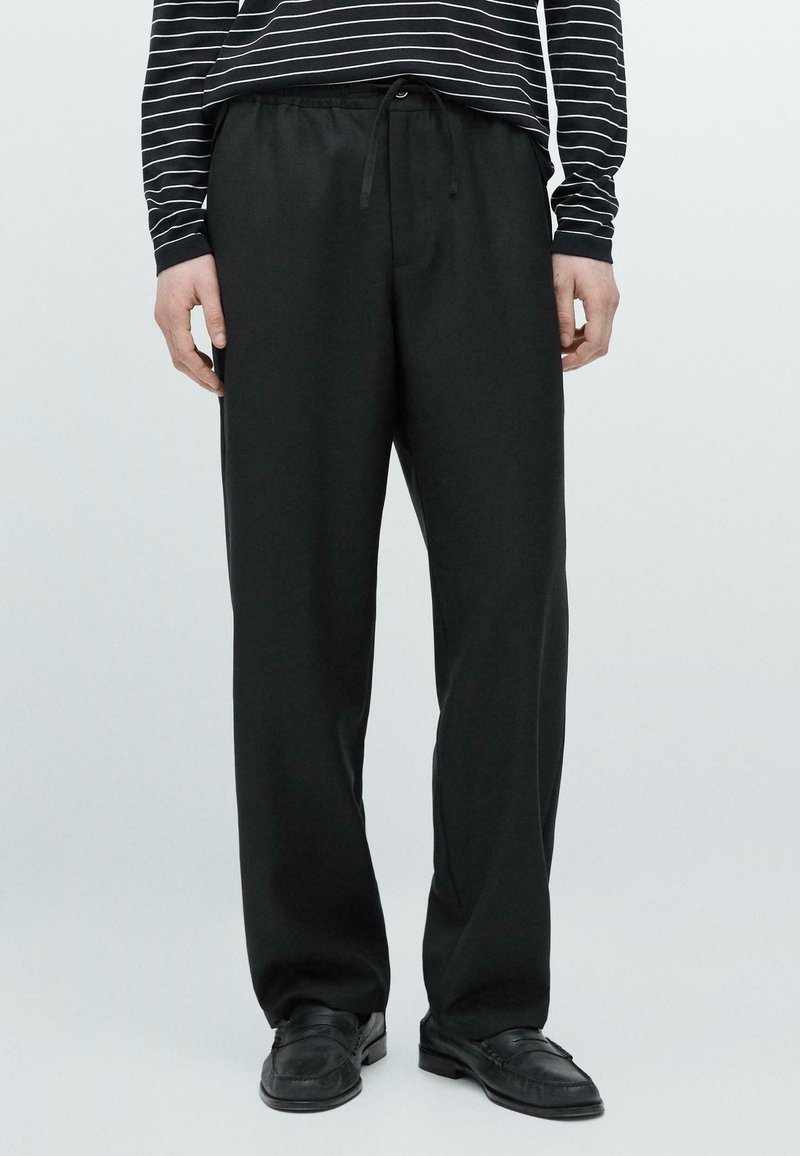 Personne portant un pantalon noir ample avec une taille à cordon, des chaussures en cuir noires et une chemise à manches longues rayée noire et blanche.