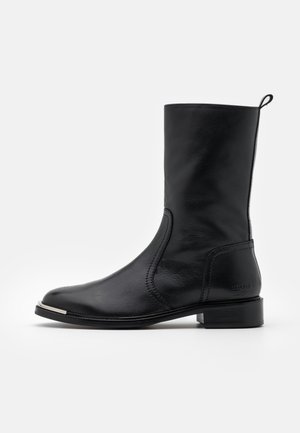Botte en cuir noir mi-mollet avec talon bas, languette pour tirer, bout arrondi, détail en métal argenté à la pointe de la semelle et marquage embossé discret sur le talon.