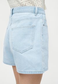 Ανοιχτό μπλε τζιν σορτς με πίσω τσέπες, φορεμένα με λευκό δαντελένιο τοπ, εμφανιζόμενα από τη πλευρά σε ανοιχτό φόντο.