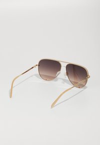 Gafas de sol aviador de metal dorado con lentes marrones degradados, varillas finas y almohadillas nasales transparentes. La montura presenta un brillo sutil.