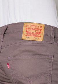 Pantalones Levi's 502 en una tela morada apagada, con una etiqueta beige con impresión roja, bolsillos traseros y costuras en contraste.