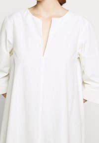 Bílé tričko s výstřihem do V, tříčtvrtečními rukávy a rovného střihu. Obsahuje jemnou žebrovanou texturu a detail středového švu na přední straně.