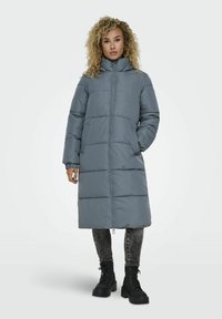 Manteau doudoune long gris avec col montant, motif matelassé horizontal, fermeture éclair frontale et poches latérales, associé à des bottines noires et un jean.