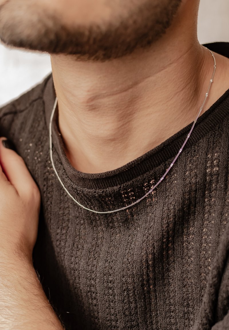 Deux colliers en argent superposés, avec des chaînes fines présentant des détails subtils, reposant sur un pull noir texturé.