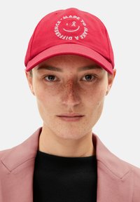 Red cap made of fabric with a curved brim. Features front embroidery of a smiley face and text: "MADE TO MAKE A DIFFERENCE."