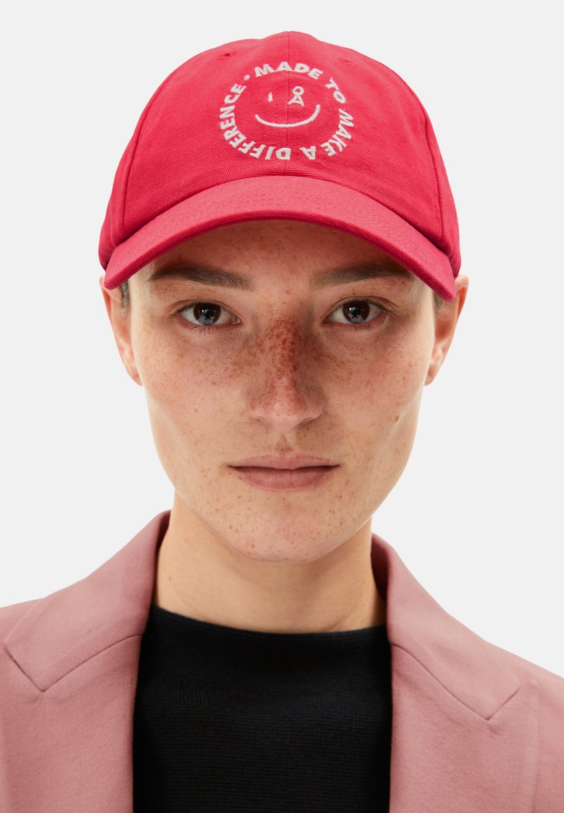 Red cap made of fabric with a curved brim. Features front embroidery of a smiley face and text: "MADE TO MAKE A DIFFERENCE."