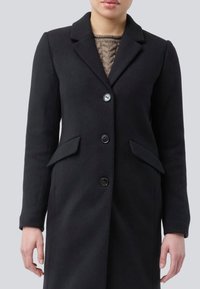 Femme portant un manteau noir à boutons par-dessus un pull beige en maille torsadée, visible de l'épaule jusqu'à la mi-cuisse sur un fond gris.