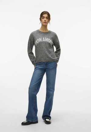 Maglione grigio con la scritta "MON AMOUR" in bianco, abbinato a jeans blu a zampa d'elefante e mocassini neri, su uno sfondo bianco semplice.