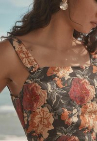 Robe fleurie avec une base noire ornée de grandes fleurs rouges et orange, encolure carrée et larges bretelles. Détail de tissu texturé visible.