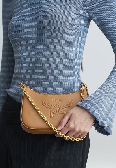 Donna che indossa un top a maniche lunghe a righe blu, tiene in mano una borsa MCM color crema con tracolla in catena dorata, abbinata a pantaloni neri.