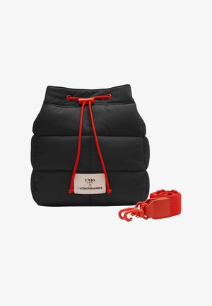 Sac seau matelassé noir à cordon avec cordons rouges et bandoulière rouge amovible, arborant une étiquette blanche avec logo sur le devant.