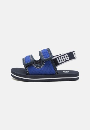Sandalia azul y negra con dos tiras ajustables y tira trasera elástica con la marca "UGG" en una suela plana con una franja blanca.