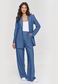 Niebieski lniany dwuczęściowy zestaw składający się z długiej marynarki i luźnych spodni, uzupełniony białą bluzką i białymi trampkami.