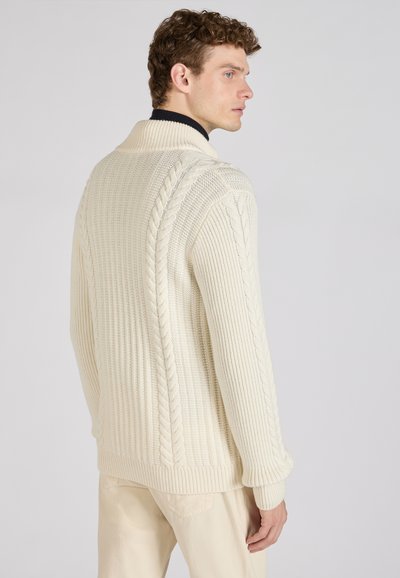 Beige gebreide cardigan met ribbelstructuur, voorzien van een sjawlkol en kabelbreipatronen langs de schouders en zijkanten.