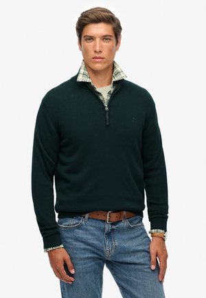 Suéter de punto verde oscuro con cuello con cremallera, usado sobre una camisa de cuadros, combinado con jeans azules y un cinturón marrón. Corte informal, textura suave.