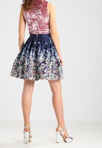 Hame floral dengan pola di atas kain biru navy, desain lipit dengan permukaan mengilap. Dipadukan dengan atasan beludru pink berbahan tekstur dan sepatu hak tinggi perak.