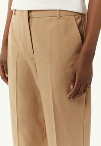 Beżowe dopasowane spodnie z gładką teksturą, plisami, klasycznym paskiem w talii i bocznymi kieszeniami. Widoczna ręka oparta na tkaninie.