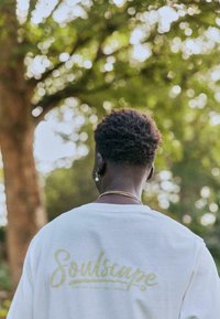 T-shirt blanc avec un graphisme vert "Soulscape", manches courtes et coupe décontractée, sur un fond flou en plein air.