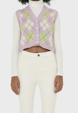 Personne portant un gilet tricoté court lavande à motifs losanges blancs et verts, boutonné, sur un col roulé blanc côtelé et un pantalon blanc.