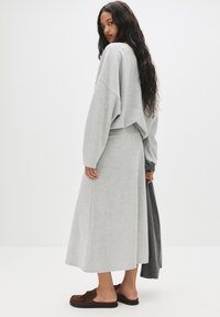Sweat-shirt gris et jupe longue assortie en tissu doux, présentant une coupe ample et des épaules dégagées ; des chaussures à enfiler marron viennent compléter la tenue.