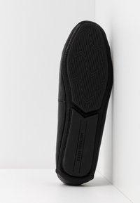 Semelle de chaussure mocassin noire avec une bande de roulement texturée et un marquage "Michael Kors" sur la section du talon, positionnée verticalement contre un coin blanc.