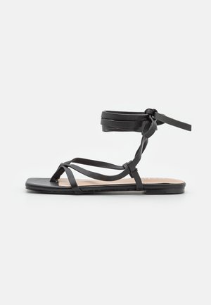 Sort flad sandal med tynde stropper, der krydser foden, og en ankelrem bundet i en sløjfe, vist på en hvid baggrund.