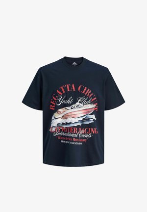 Garçon souriant debout, portant un t-shirt graphique noir du yacht club, un short en denim clair, des chaussettes blanches et des chaussures beige à lacets sur un fond uni.