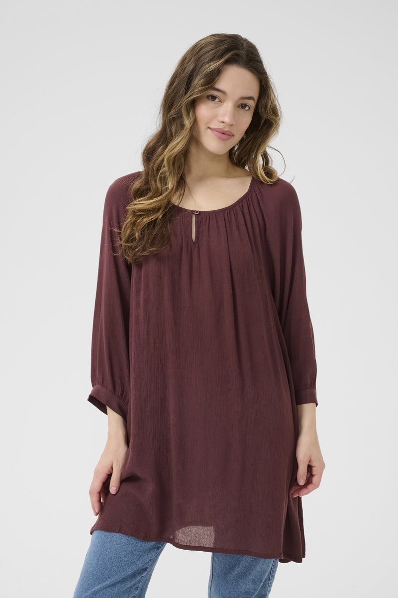 Tunique bordeaux à manches longues, avec un décolleté froncé et un détail subtil en forme de trou de clé. Fabriquée dans un tissu léger et texturé.