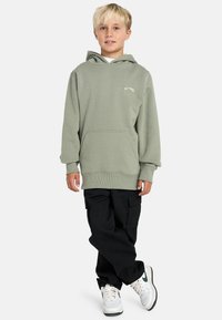 Felpa verde con cappuccio e tasca frontale, dotata di polsini e orlo a coste, abbinata a pantaloni cargo neri e sneakers bianche.
