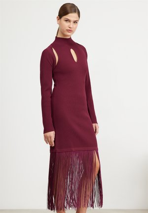 Robe bordeaux à côtes avec manches longues, détails découpés aux épaules et ourlet à franges atteignant le mollet. Texture lisse et style ajusté.
