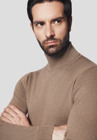 Brązowy sweter z golfem wykonany z prążkowanej dzianiny, o dopasowanym kroju, z długimi rękawami i teksturowanym wykończeniem. Model pozuje z założonymi rękami.