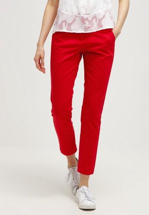 Vrouw draagt felrode, nauwsluitende enkellange broek, witte kanten top en witte sneakers, lopend met één hand in de zak.