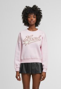 Felpa rosa con scollatura arrotondata, caratterizzata da un grande logo ricamato e un design di stelle. Tessuto morbido e testurizzato con polsini a costine. Gonna nera plissettata.