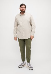 Camisa abotonada de color beige claro, pantalones verdes ajustados y zapatillas deportivas grises. La camisa tiene mangas largas y un collar clásico.