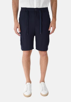 Uomo che indossa pantaloncini cargo blu navy con coulisse e bottone, scarpe da ginnastica bianche e camicia bianca a maniche corte, in piedi su uno sfondo semplice.