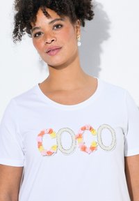 Camiseta blanca con la palabra "COCO" adornada con coloridos apliques florales y acentos de cuentas, escote redondo y mangas cortas.