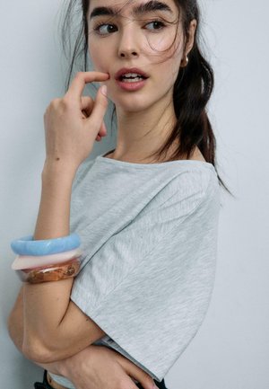 Mlada ženska s temnimi lasmi v sivi majici se dotika obraza, na levi roki pa nosi tri masivne zapestnice v modri, beli in jantarni barvi.